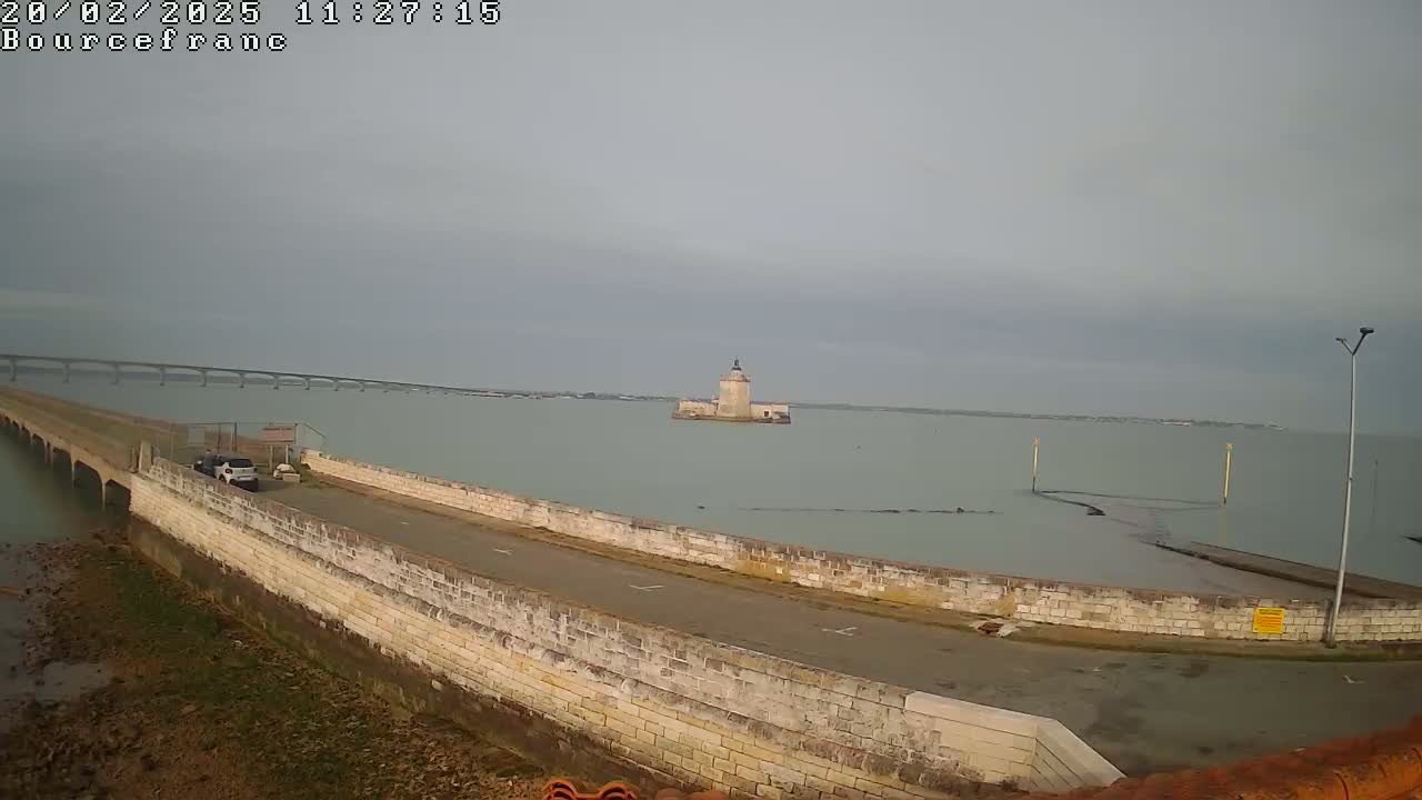 A stone causeway leads to a small island with a tower in a calm, overcast body of water, with a long bridge visible in the distance.