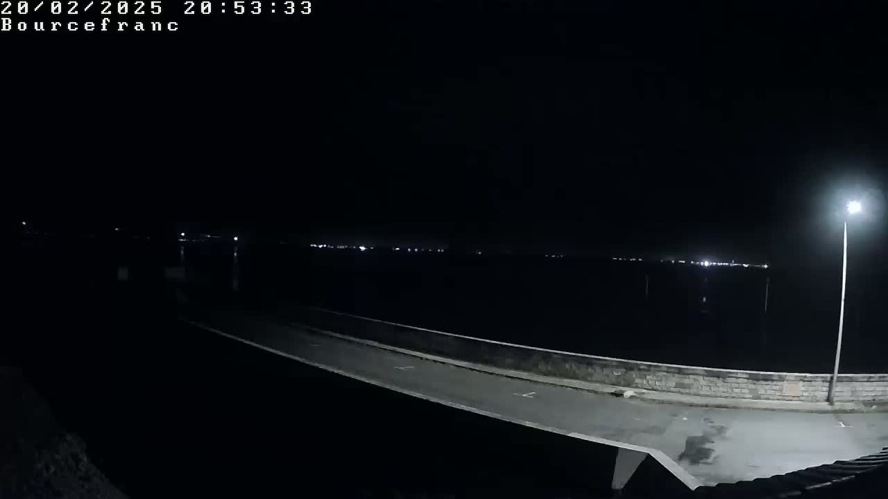 A dark, nighttime scene shows a paved roadway and low wall overlooking a body of water with distant lights.