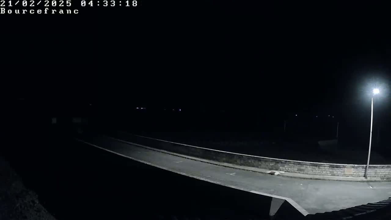 A nighttime view of a dark, empty road illuminated by a single streetlight, with a low stone wall alongside it.