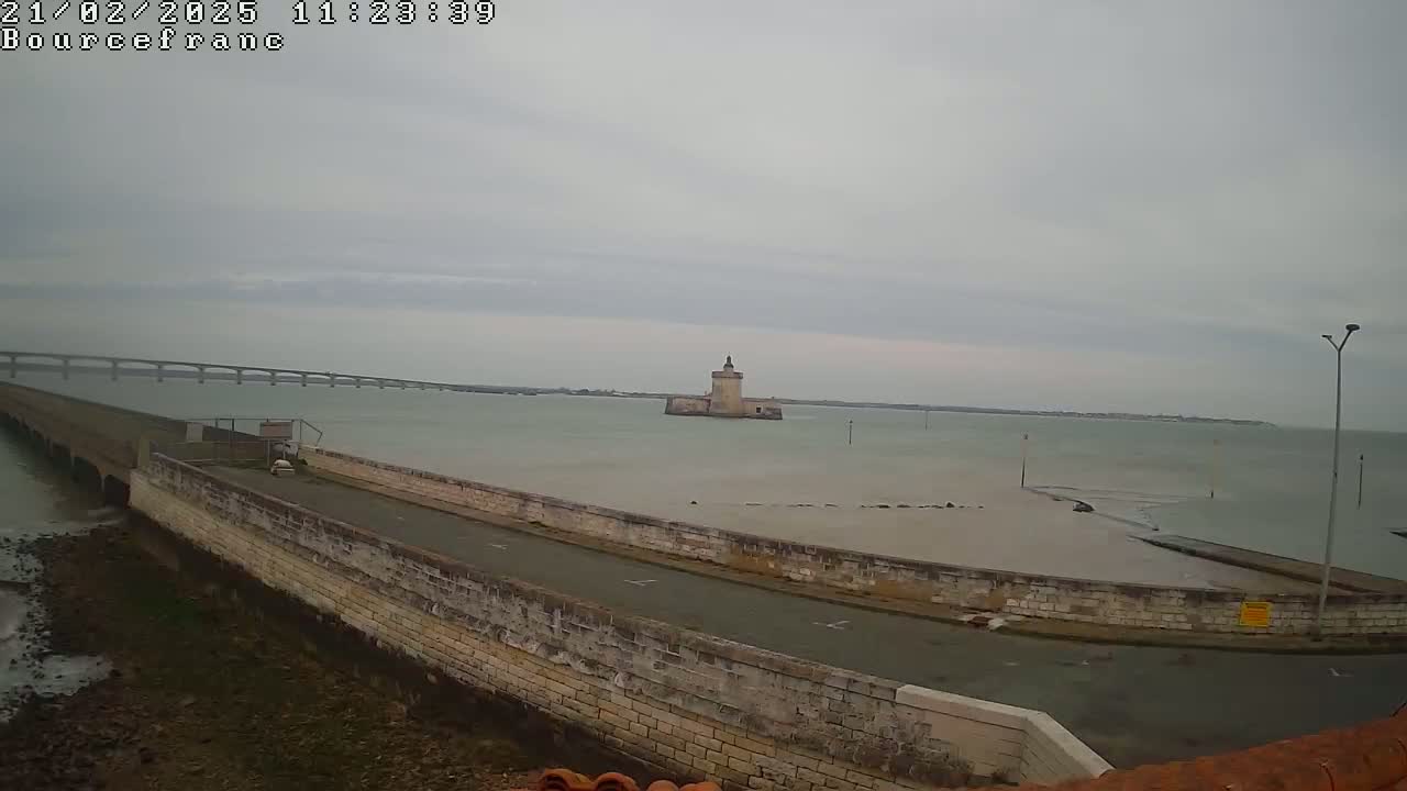 A stone wall and road curve around a shallow, calm body of water under an overcast sky, with a distant bridge and a stone tower visible in the background.