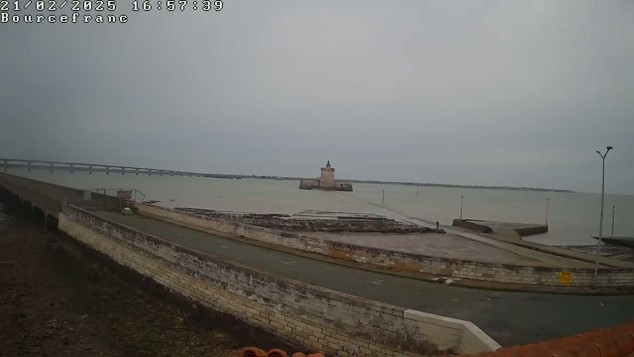 A stone structure and roadway extend into a calm, overcast body of water, with a distant bridge visible in the background.