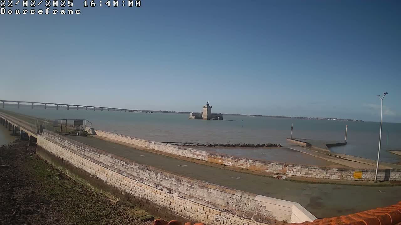 A stone wall and roadway curve around a tidal area, leading to a distant stone tower and a long bridge under a clear blue sky.
