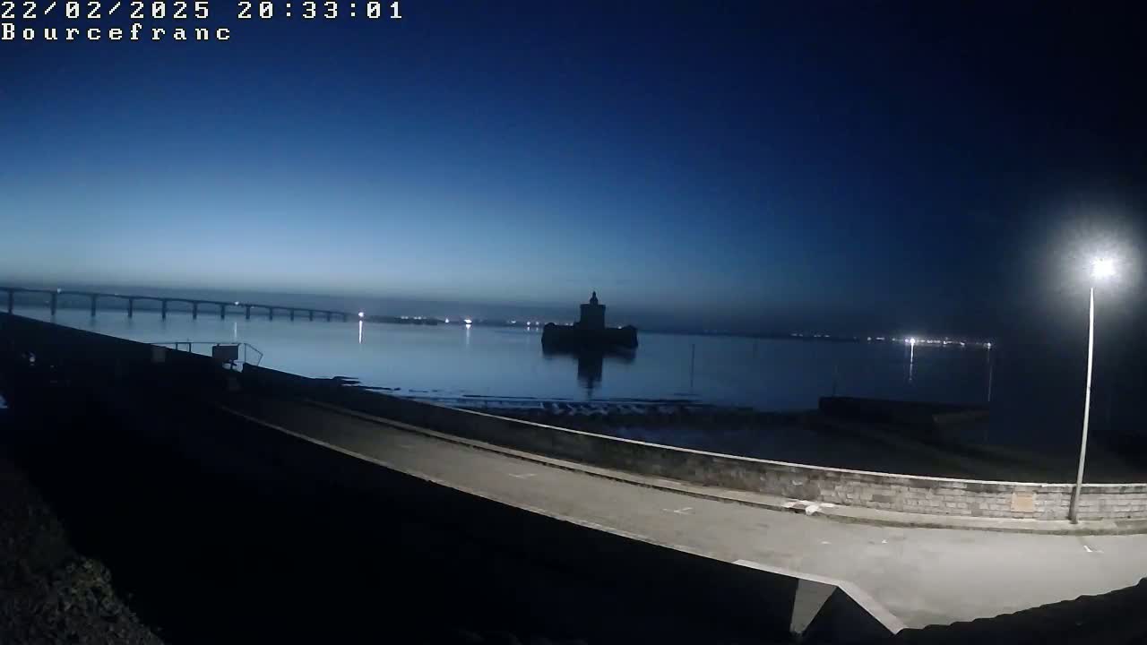 A dark, calm body of water stretches out under a twilight sky, featuring a distant bridge, a solitary castle-like structure on an islet, and a paved road in the foreground lit by a streetlamp.