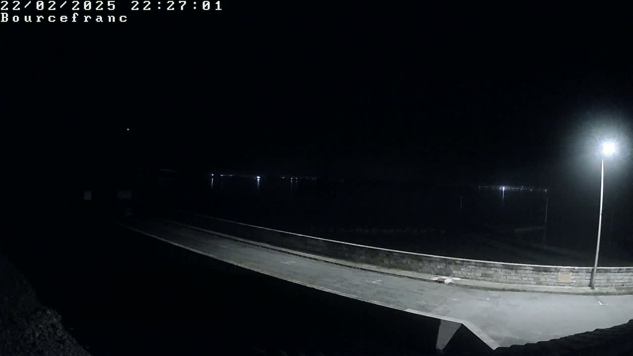 A nighttime view shows a dimly lit road and a low stone wall overlooking a dark body of water under a clear, dark sky.