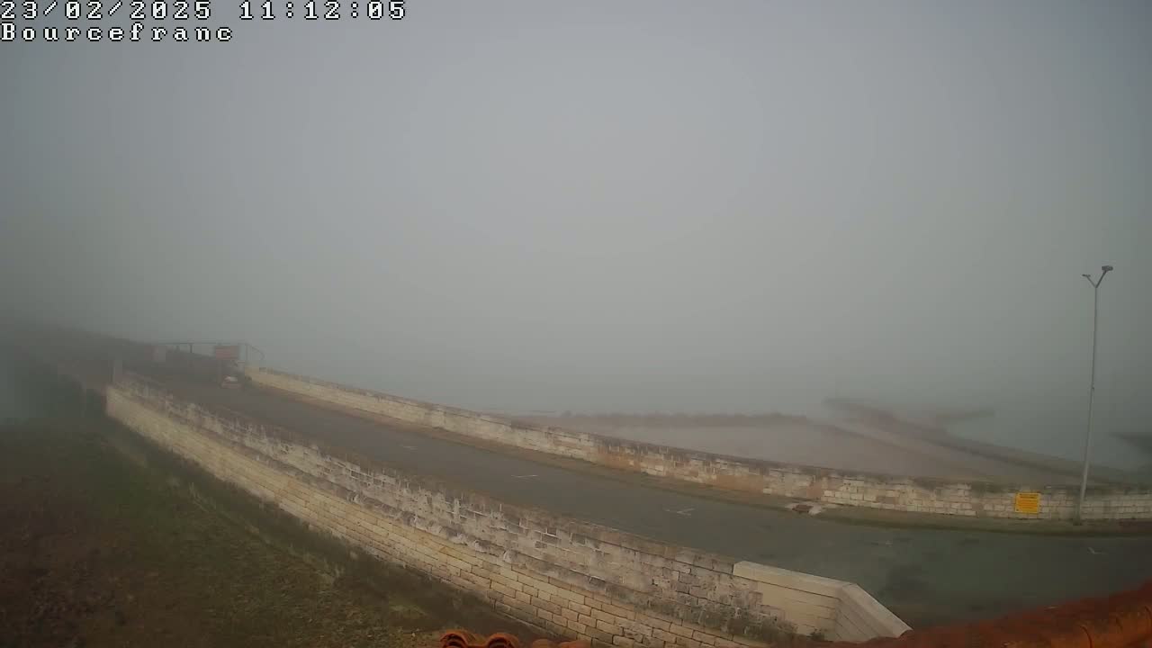 A foggy view shows a paved road along a low stone wall overlooking a body of water shrouded in mist.