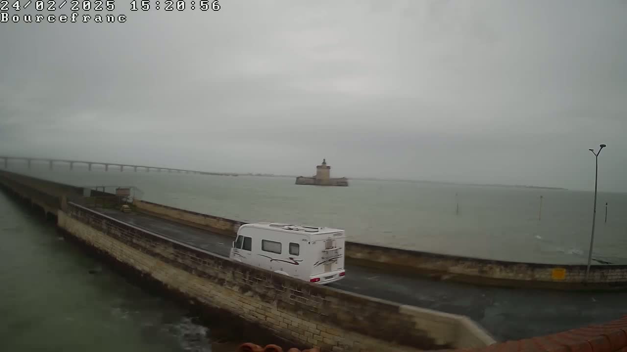 A white camper van drives along a seaside road on an overcast day, with a distant bridge and a stone tower visible across the water.