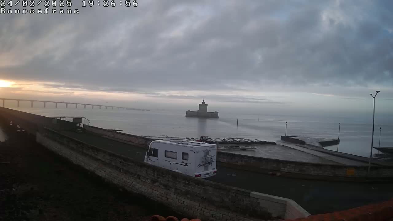 A white camper van is parked near a stone wall overlooking a calm body of water, a distant bridge, and a stone tower under a cloudy, sunrise sky.