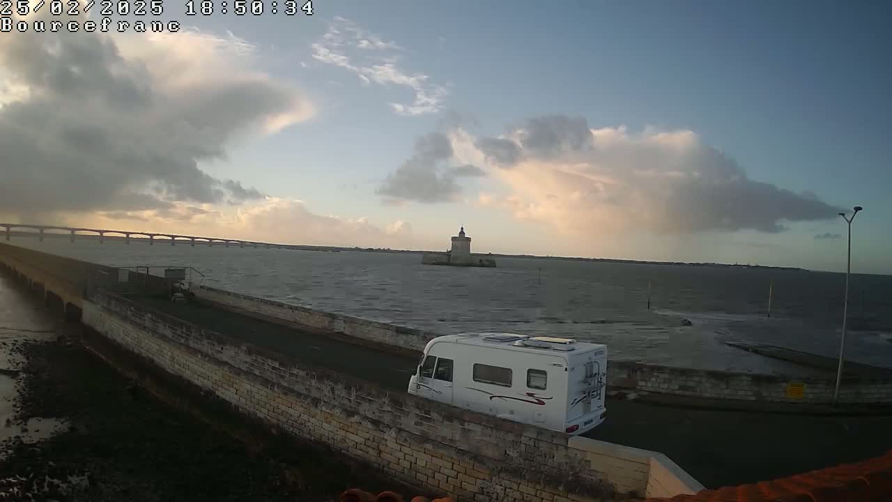A campervan drives along a stone wall overlooking a body of water with a distant bridge and a small stone tower under a partly cloudy sky.
