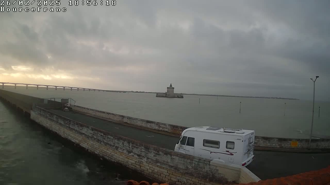 A white camper van drives across a narrow causeway toward a stone tower in the distance, under an overcast sky on a somewhat choppy body of water.