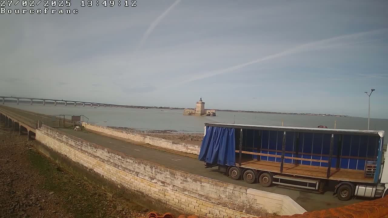 A flatbed truck is parked near a stone wall overlooking a calm body of water, a distant bridge, and a small stone tower on an island under a mostly clear sky.