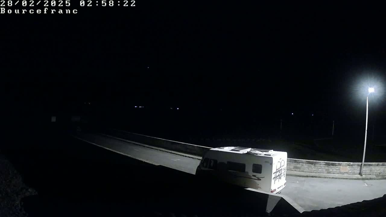 A white camper van is parked at night on a paved road next to a low stone wall, illuminated by a single streetlight.