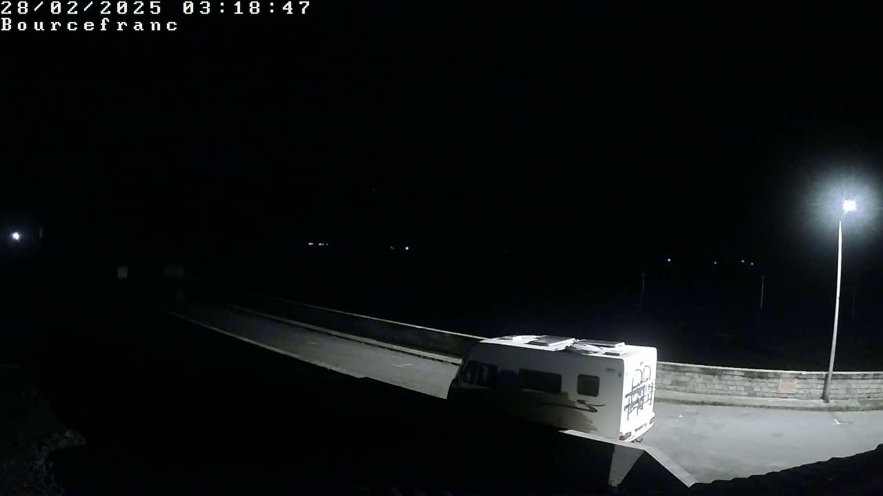 A white camper van is parked at night on a paved area next to a low wall, illuminated by several streetlights.