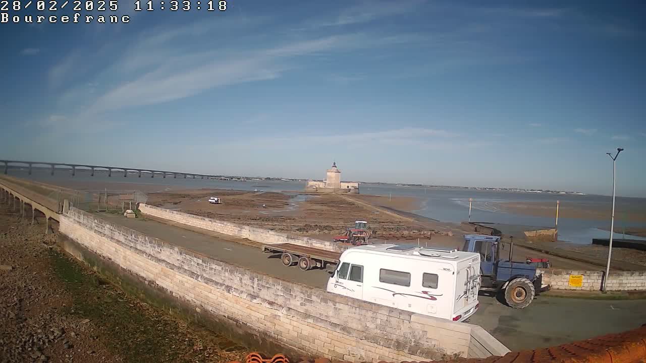A white camper van and a tractor are parked near a stone wall overlooking a tidal flat, a distant bridge, and a stone tower.
