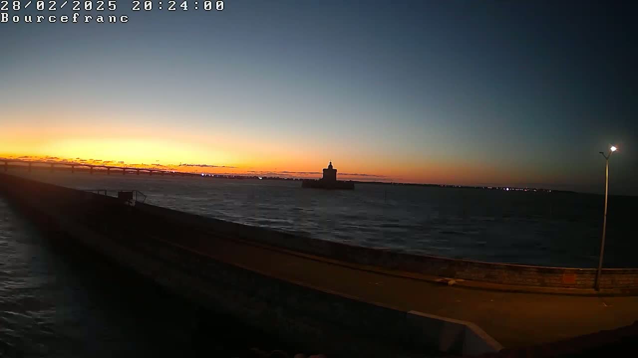 A sunset over a body of water, showing a distant bridge and a tower in the water, with a long, dark road or embankment in the foreground.