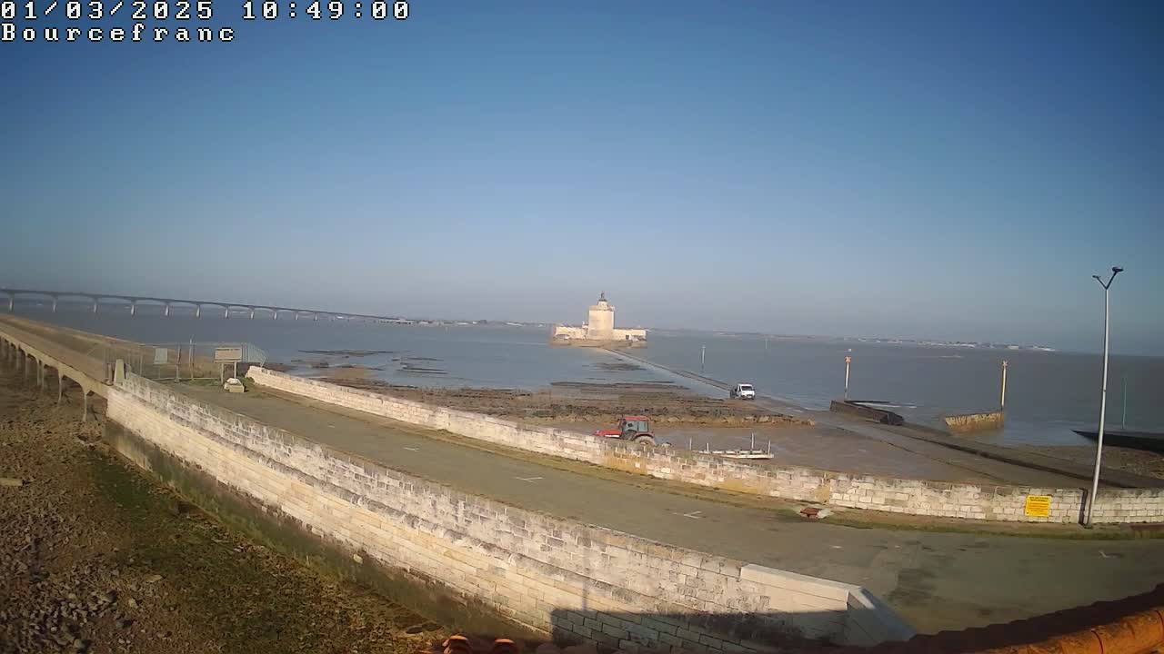 A stone causeway leads to a small island with a stone tower, a bridge is visible in the distance across a calm body of water under a clear sky.