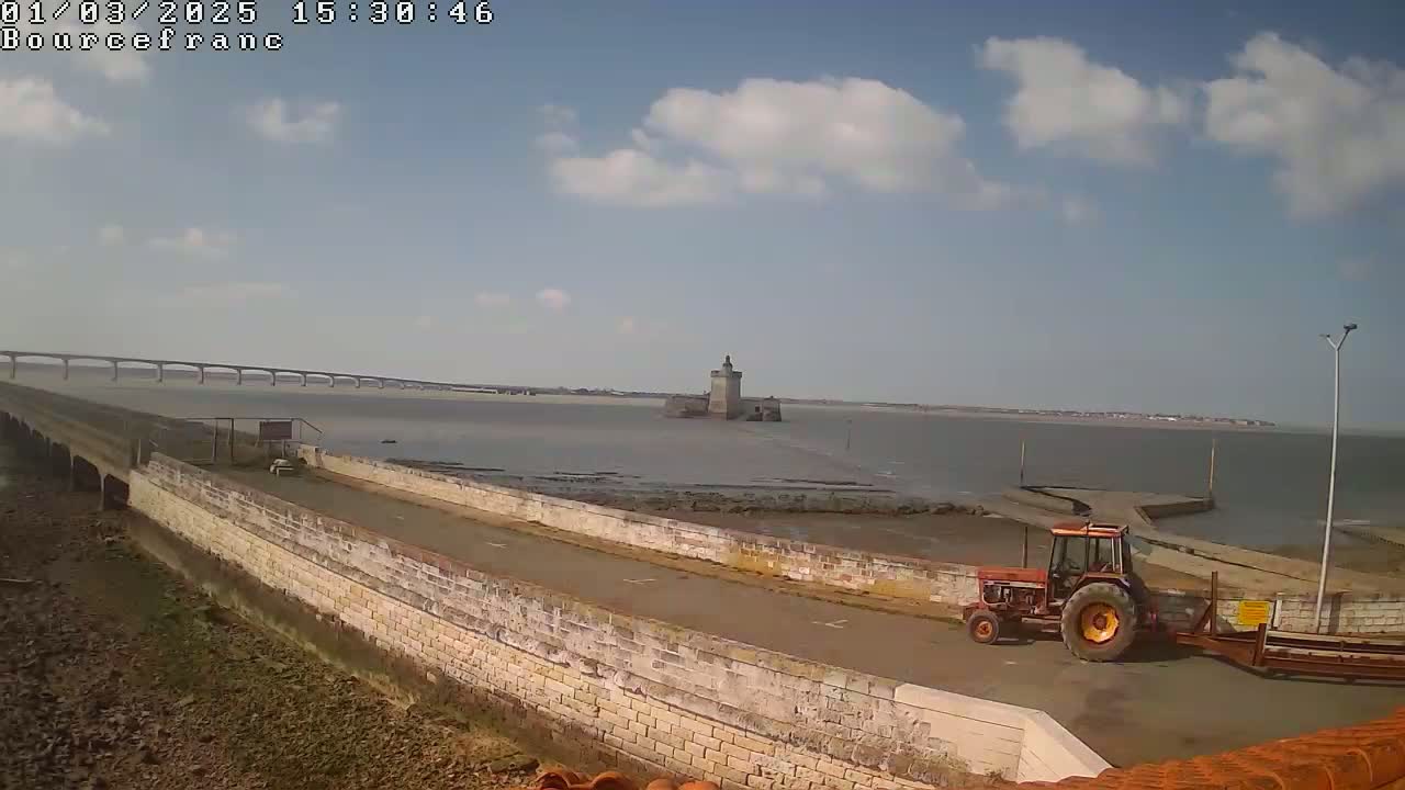 A tractor is parked near a stone wall overlooking a partly exposed bay, a distant bridge, and a stone tower under a partly cloudy sky.