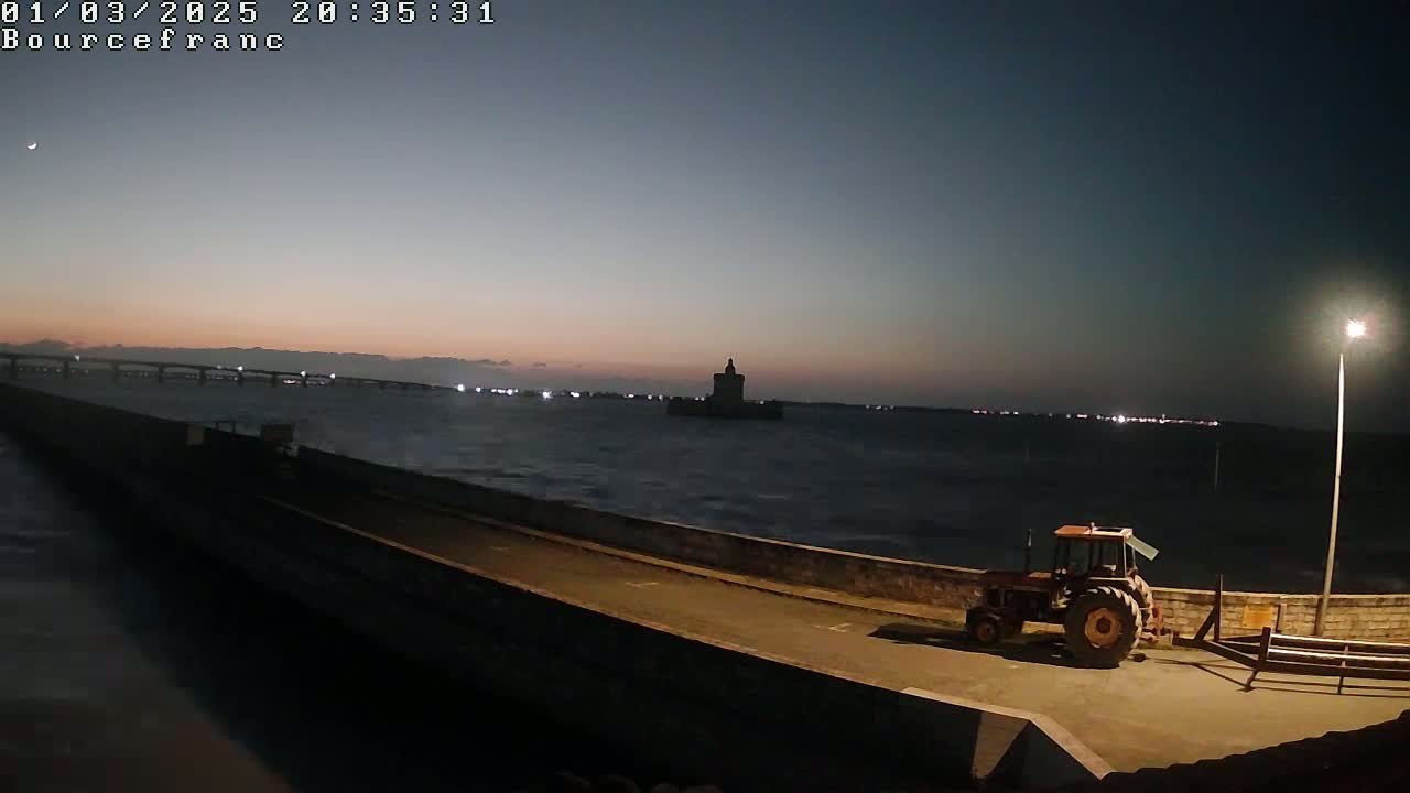 A tractor sits on a paved path next to a wall overlooking a dark, choppy body of water, with a distant bridge and tower visible under a twilight sky.