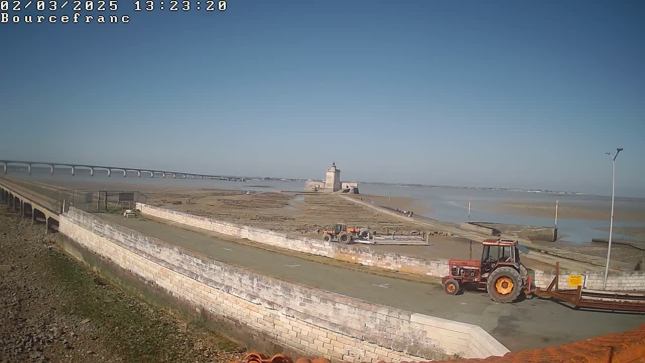 A long stone causeway leads to a tower on a tidal flat, with a large bridge visible in the distance under a clear blue sky.