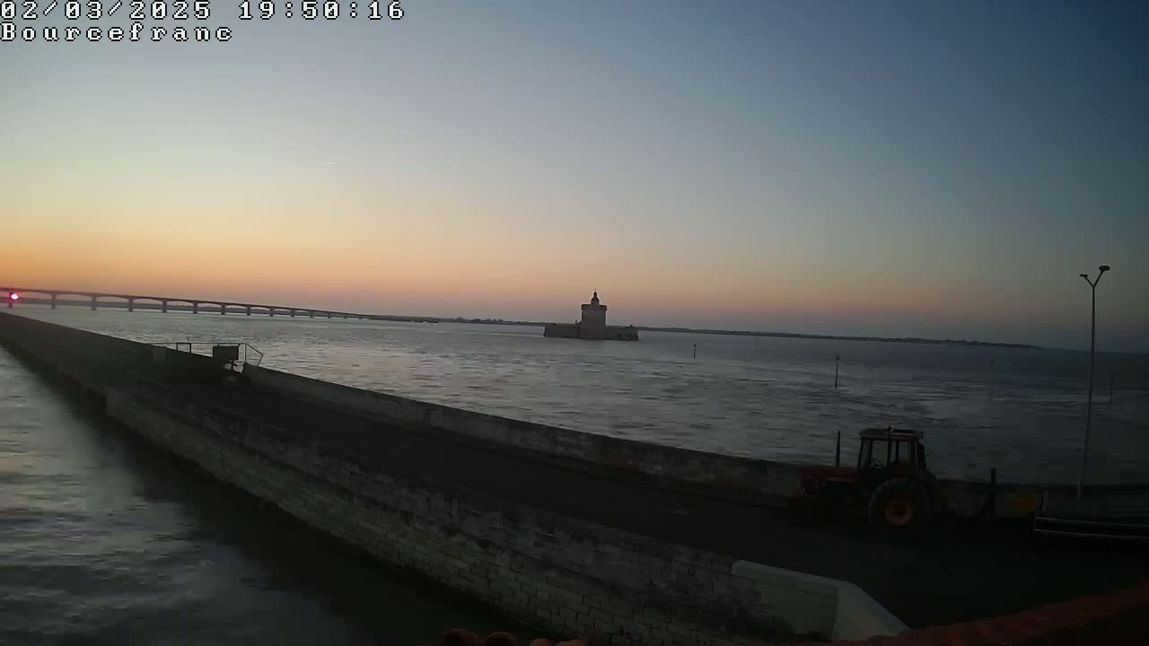 A long stone wall extends into a calm body of water at sunset, with a distant bridge, a small stone tower, and a tractor visible.