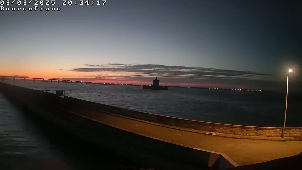 A dark silhouette of a tower is visible in the distance across a body of water, with a long bridge and a pink sunset in the background.