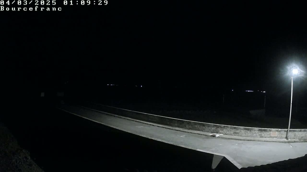 A dark, nighttime scene shows a low-lit roadway over a low wall, illuminated by a single streetlamp.