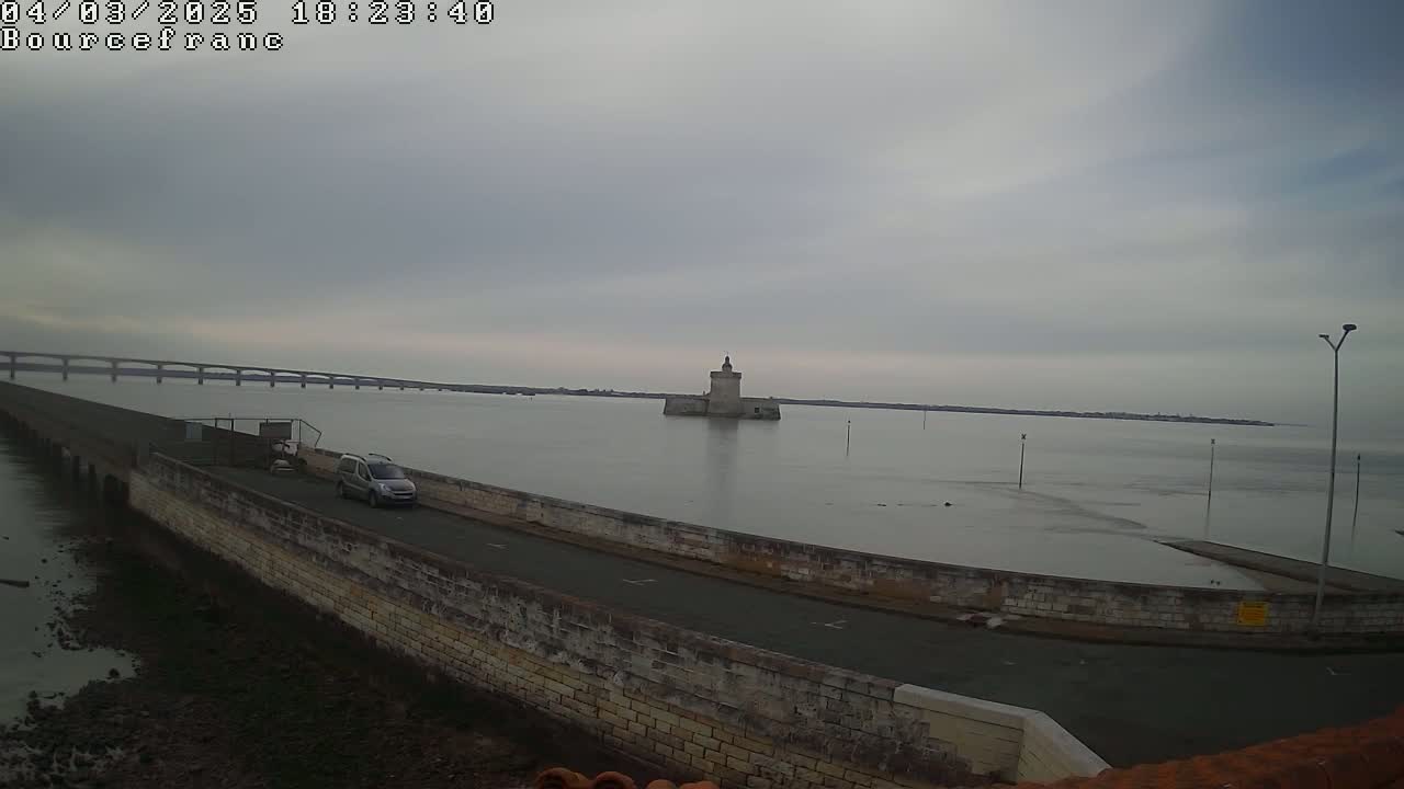 A stone wall curves around a roadway overlooking calm water, a distant bridge, and a small stone tower under an overcast sky.