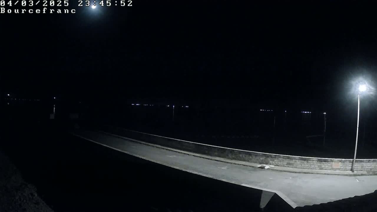 A nighttime view of a paved road leading to a low wall, illuminated by a single bright lamppost, with distant faint lights visible in the darkness.