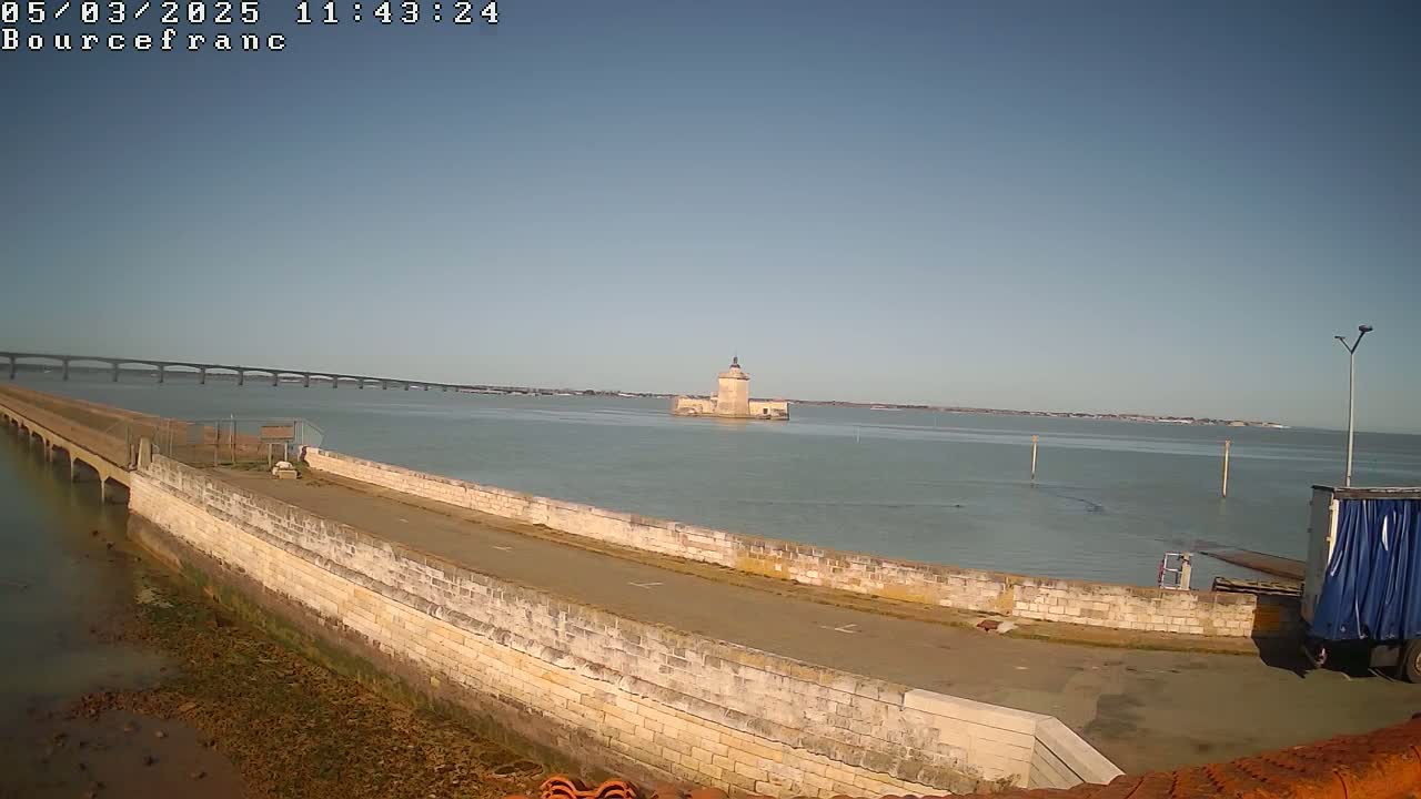 A stone wall and causeway extend into a calm body of water towards a distant tower, with a long bridge visible in the background under a clear sky.