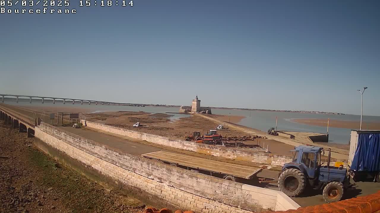 A stone causeway leads to a tidal island featuring a stone tower, tractors, and a flatbed trailer, all under a clear blue sky.