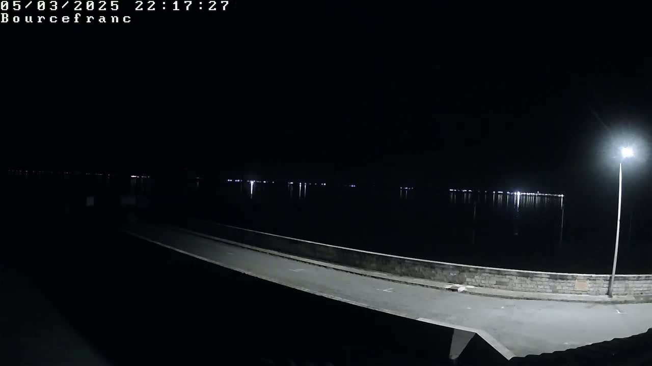 A nighttime view of a paved area next to a calm body of water reflecting distant lights under a dark sky.