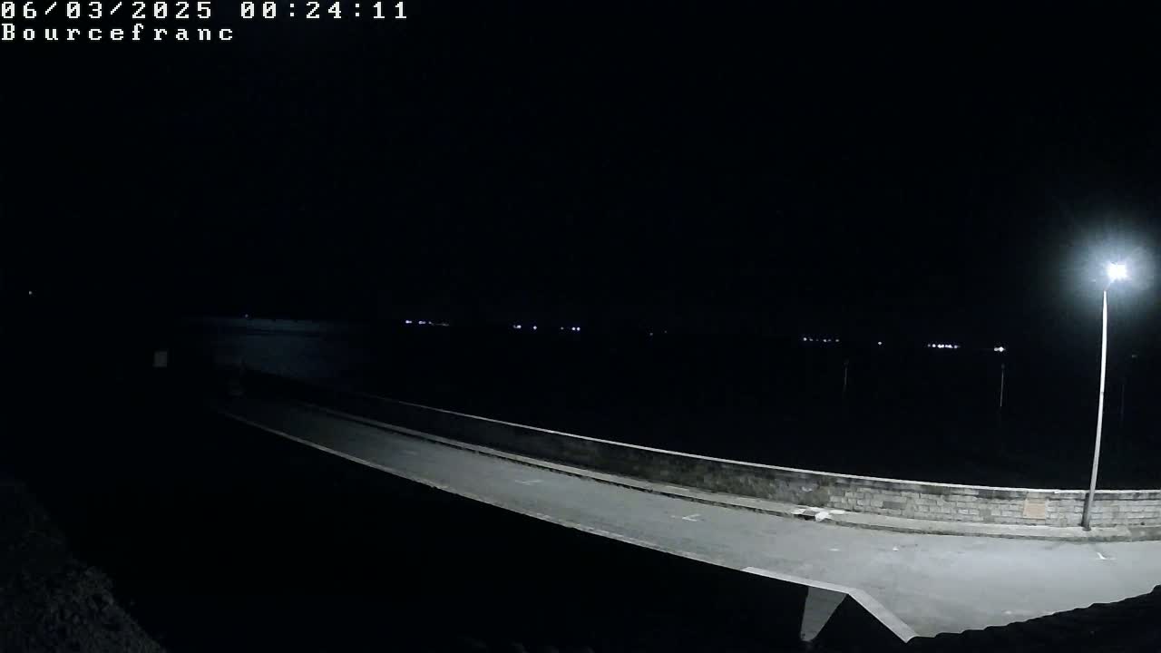 A dark, nighttime scene shows a paved roadway on a low wall overlooking a body of water, illuminated by a single lamppost.