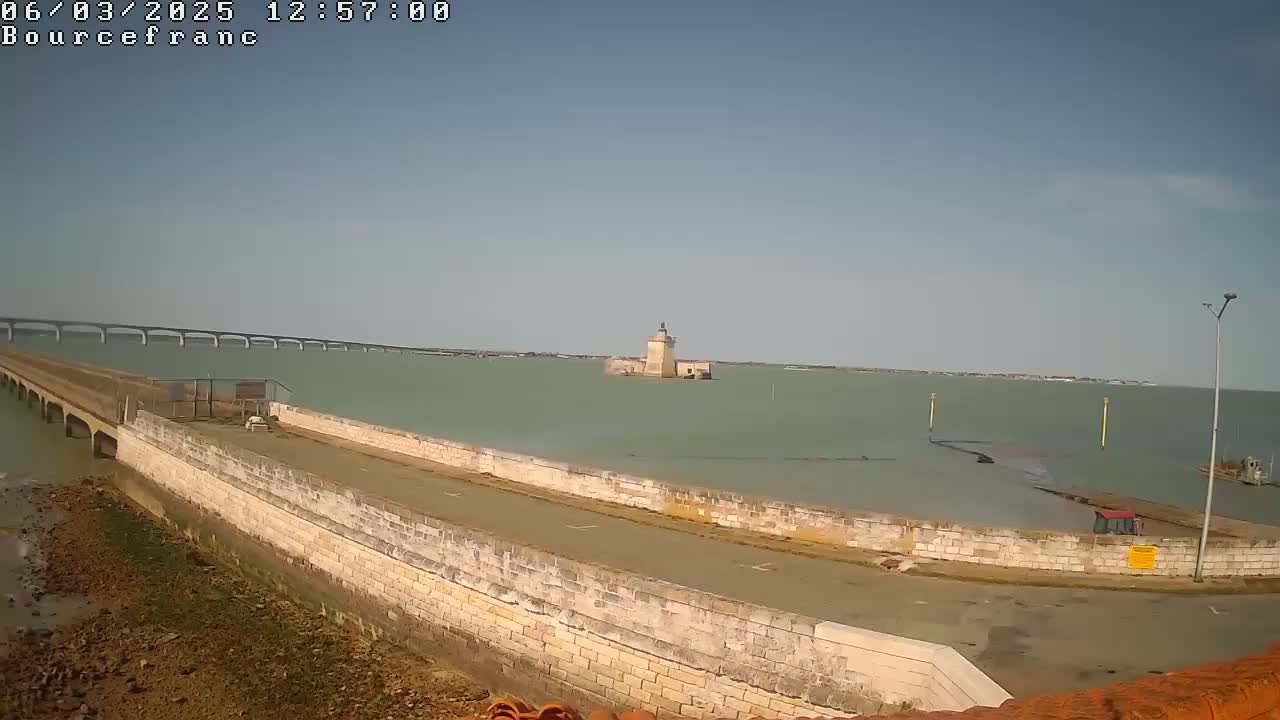A stone causeway leads to a small island with a stone tower, a long bridge is visible in the distance across calm, greenish water under a clear sky.