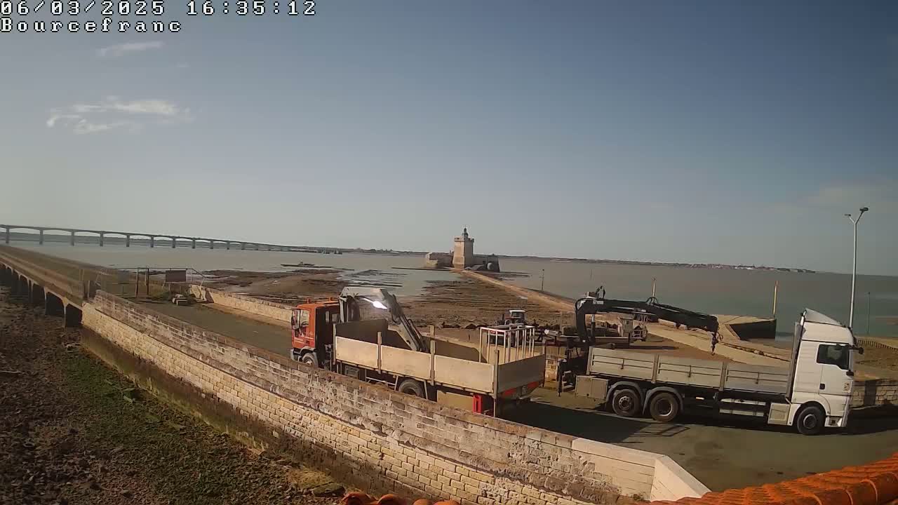 Two trucks with cranes are parked on a stone causeway near a stone fort and a long bridge under a mostly clear sky.