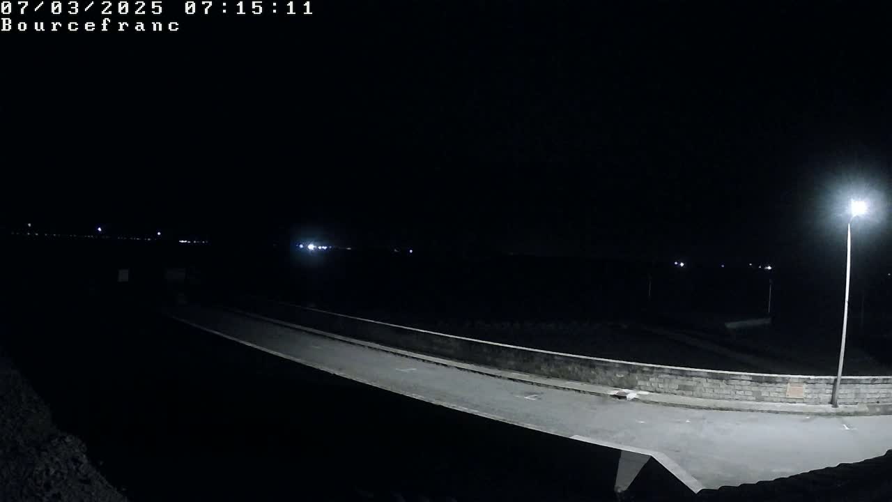 A nighttime view of a dark, empty road and bridge illuminated by a single streetlight, with distant lights visible in the background.