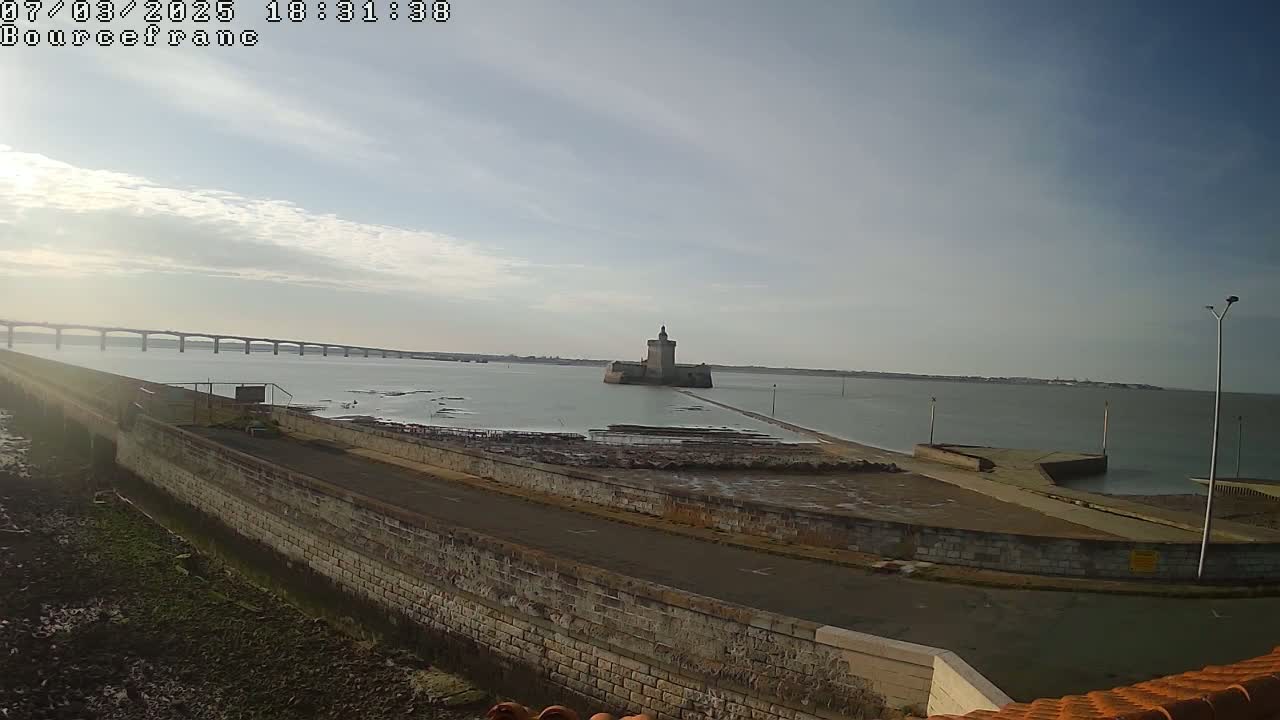 A stone seawall leads to a small island fortress, with a long bridge visible in the distance under a mostly clear sky.