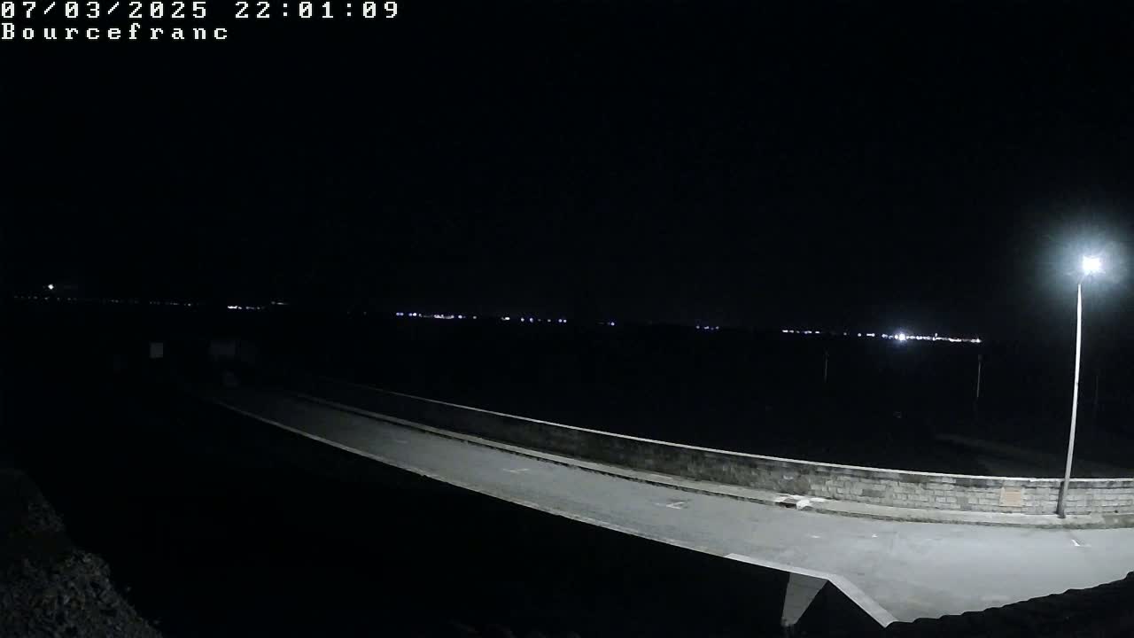 A nighttime view of a paved road leading to a low stone wall, beyond which distant lights are visible under a clear, dark sky.