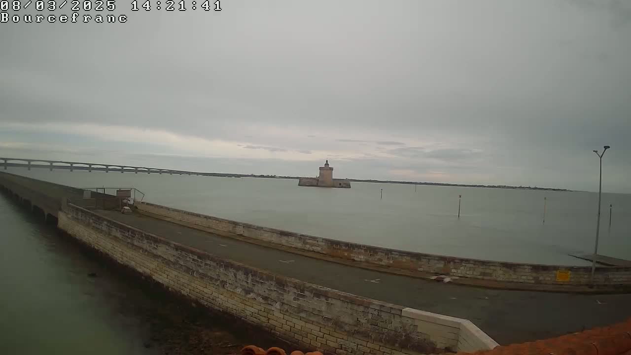 A stone tower sits in calm, gray water under an overcast sky, with a long bridge visible in the distance.