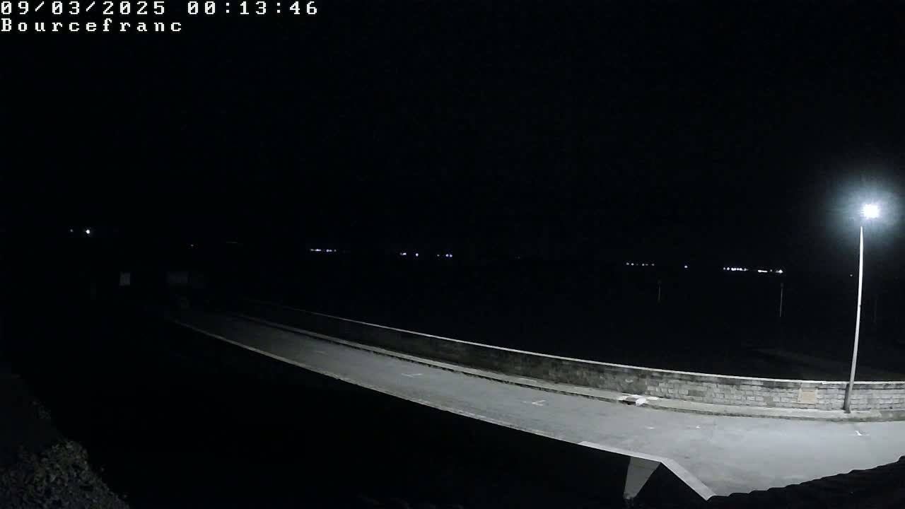 A dark, nighttime scene shows an empty roadway lit by a single lamppost extending from a low stone wall overlooking a dark body of water.