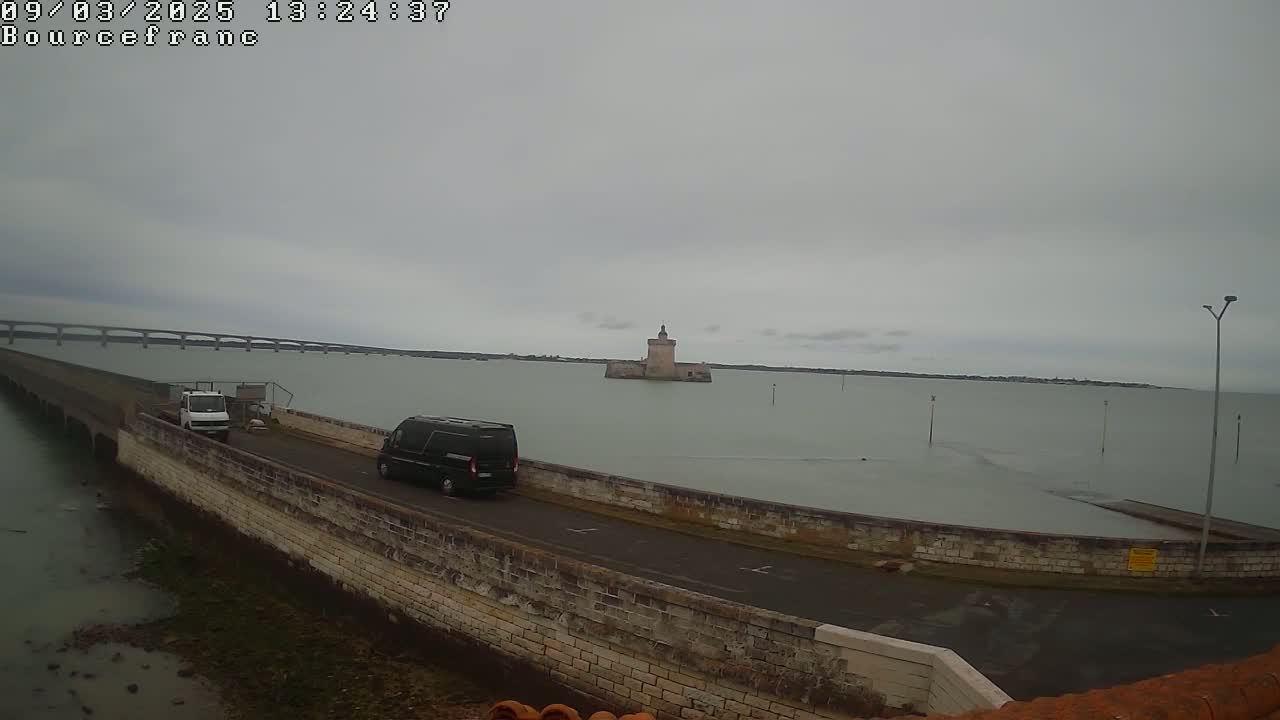 A long, low stone wall extends into calm, gray water towards a distant stone tower, with two vehicles and a bridge visible in the background under an overcast sky.