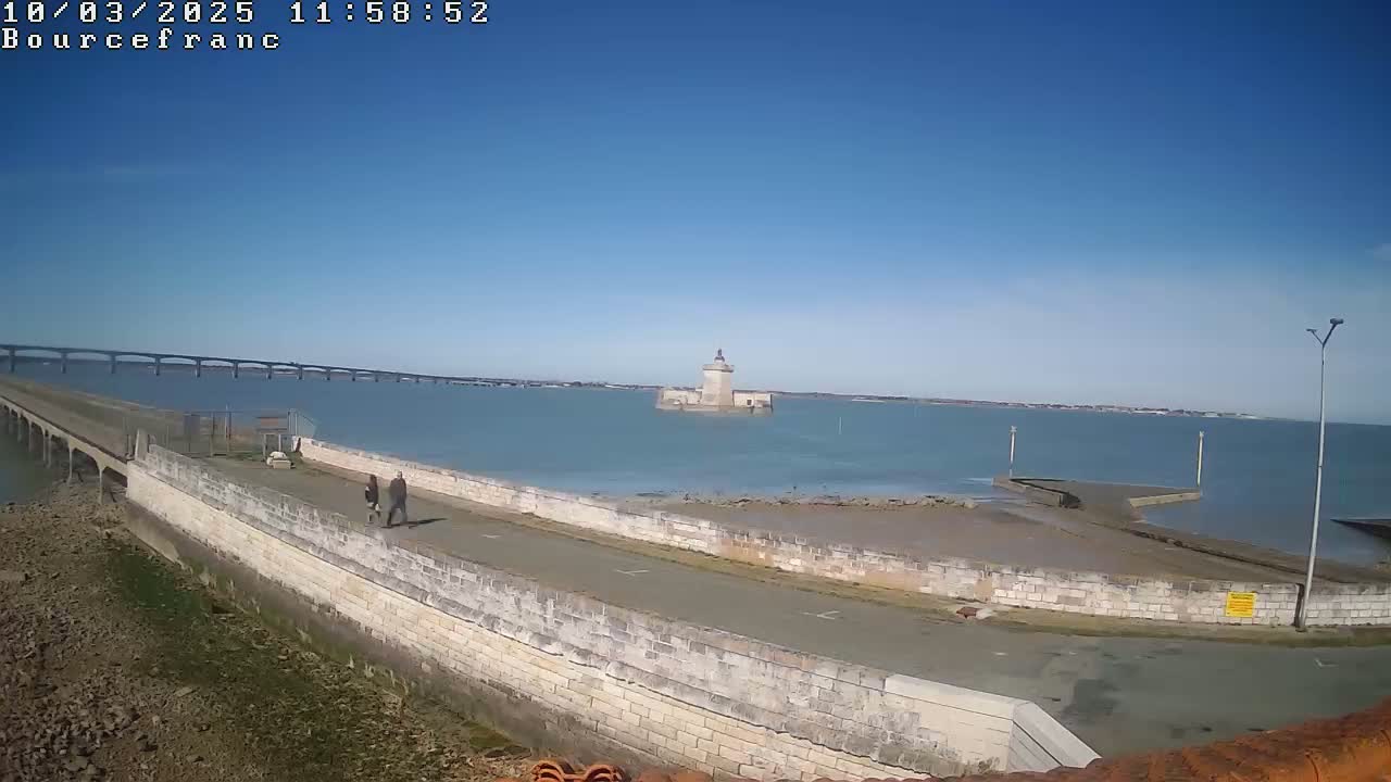 A long stone walkway extends from a viewpoint toward a stone tower in the water, with a bridge visible in the distance under a clear blue sky.