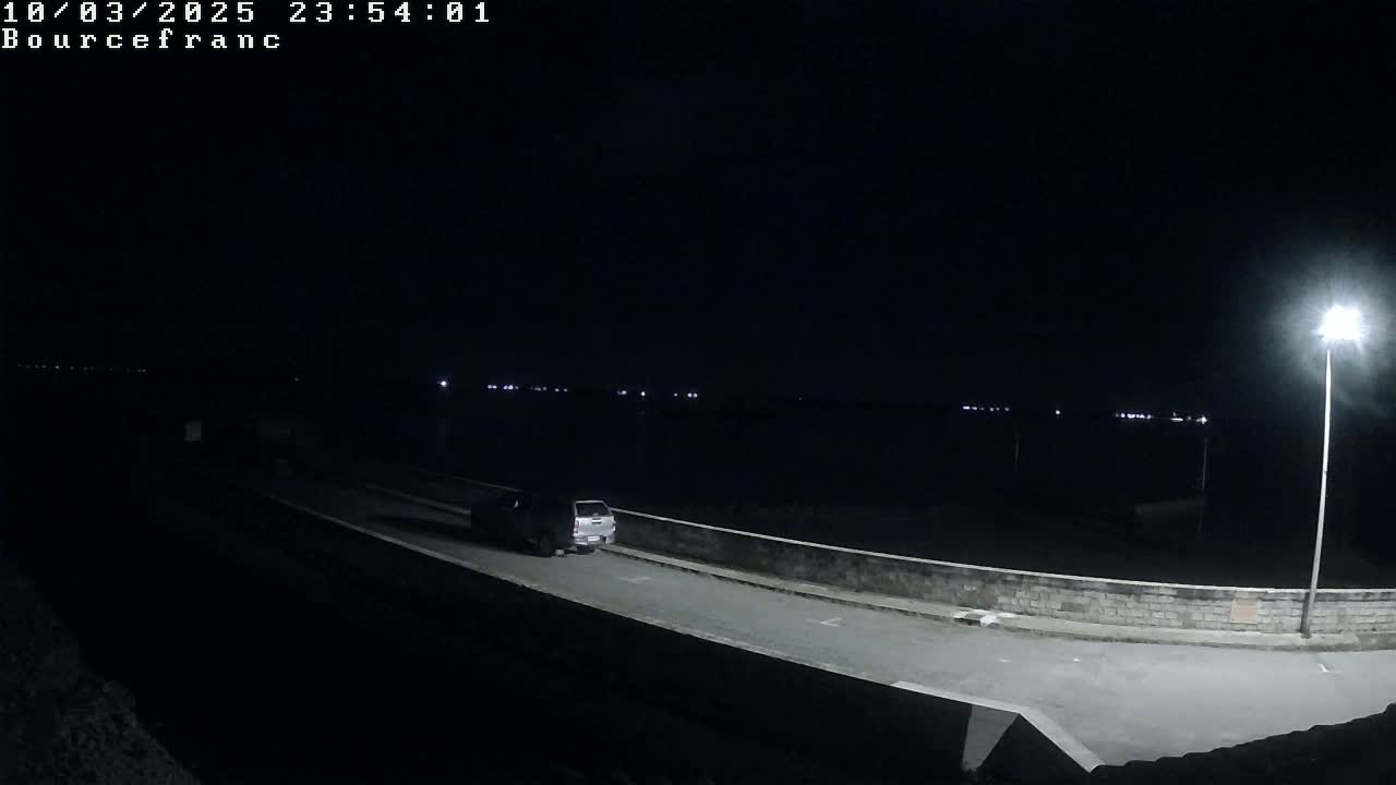 A silver pickup truck drives on a dark road at night, illuminated by a single streetlight, past a low stone wall.