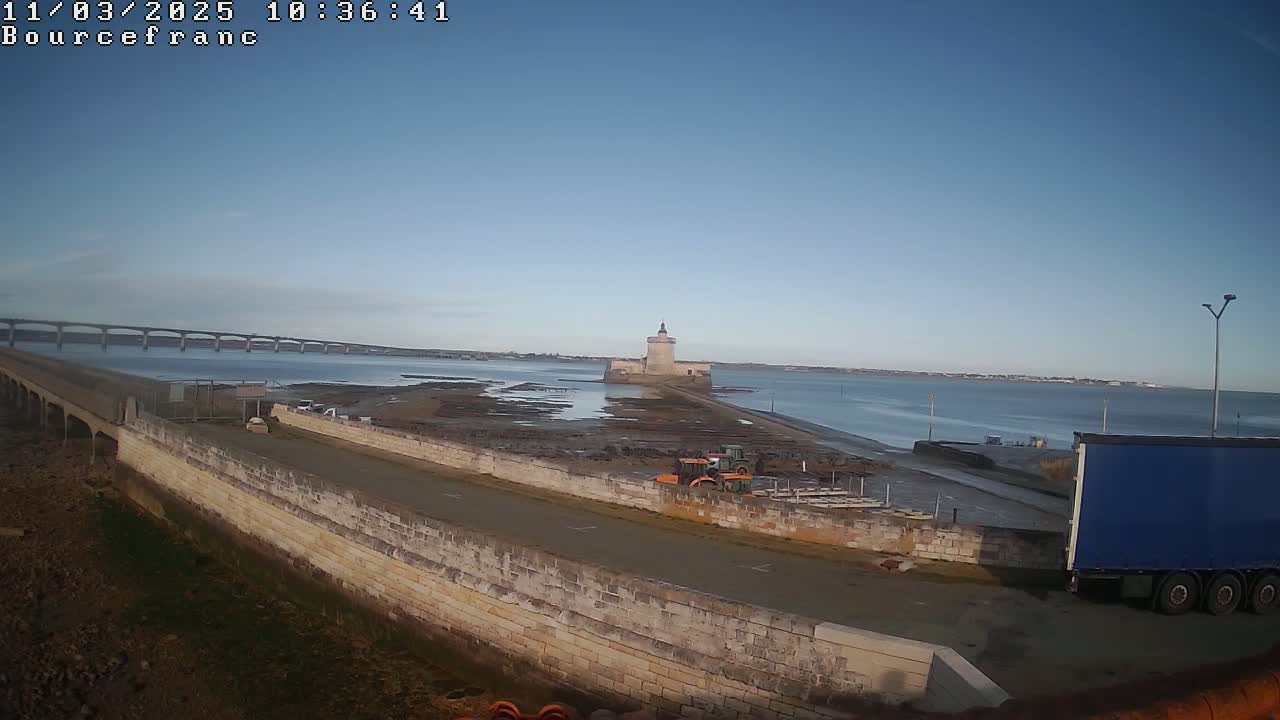 A stone causeway extends to a small, fortified tower on a calm, clear day, with a long bridge visible in the background.