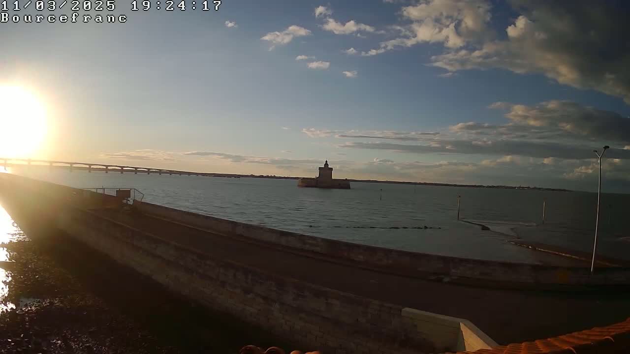 A distant stone tower sits in calm water under a partly cloudy sunset sky, with a bridge visible in the background.