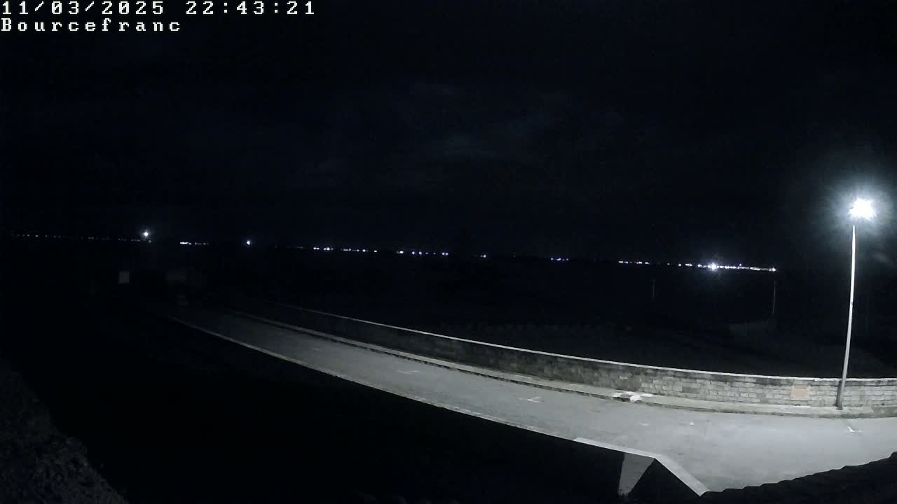 A nighttime view of a low-lit roadway with a low stone wall on either side, extending towards a distant horizon with faint lights.