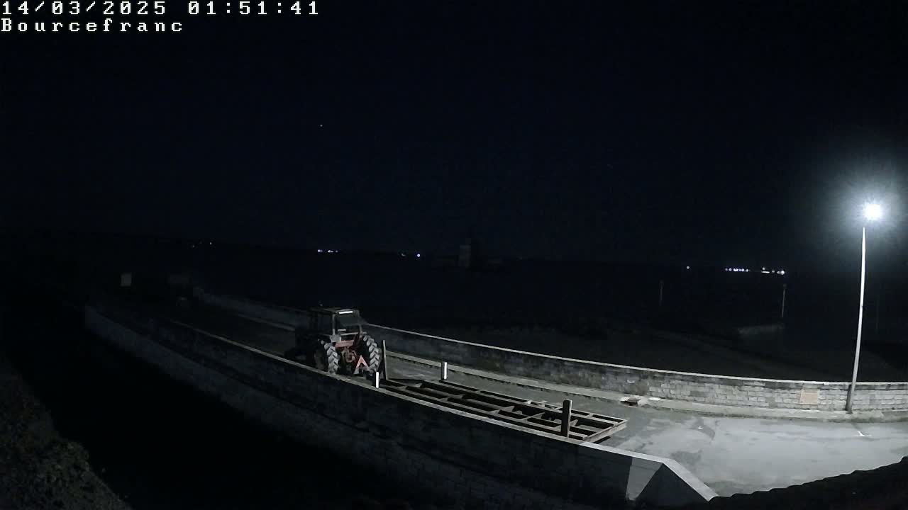A tractor sits on a dark, paved area next to a low wall overlooking a body of water at night under a light pole.