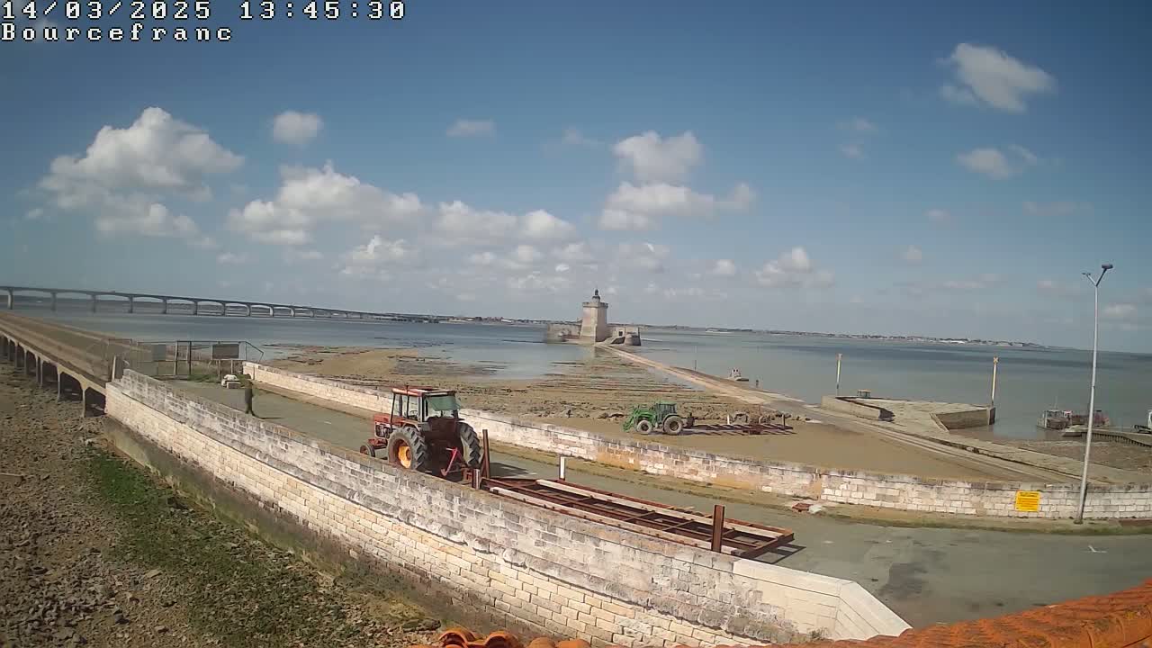 Under a partly cloudy sky, two tractors are parked near a stone wall and a tidal causeway leading to a tower, with a bridge visible in the distance.