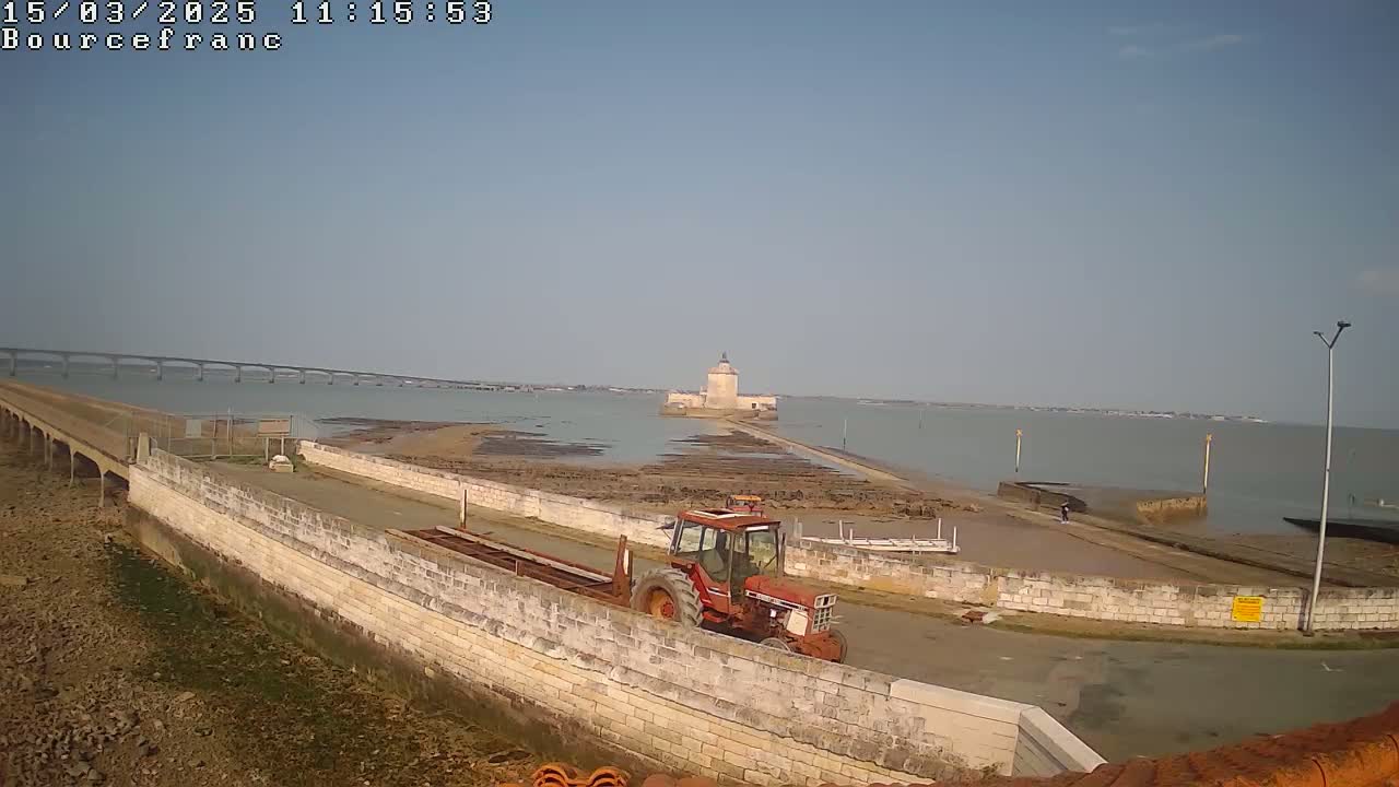 A red tractor sits on a stone causeway leading to a stone tower in the water, under a clear sky, with a bridge visible in the distance.