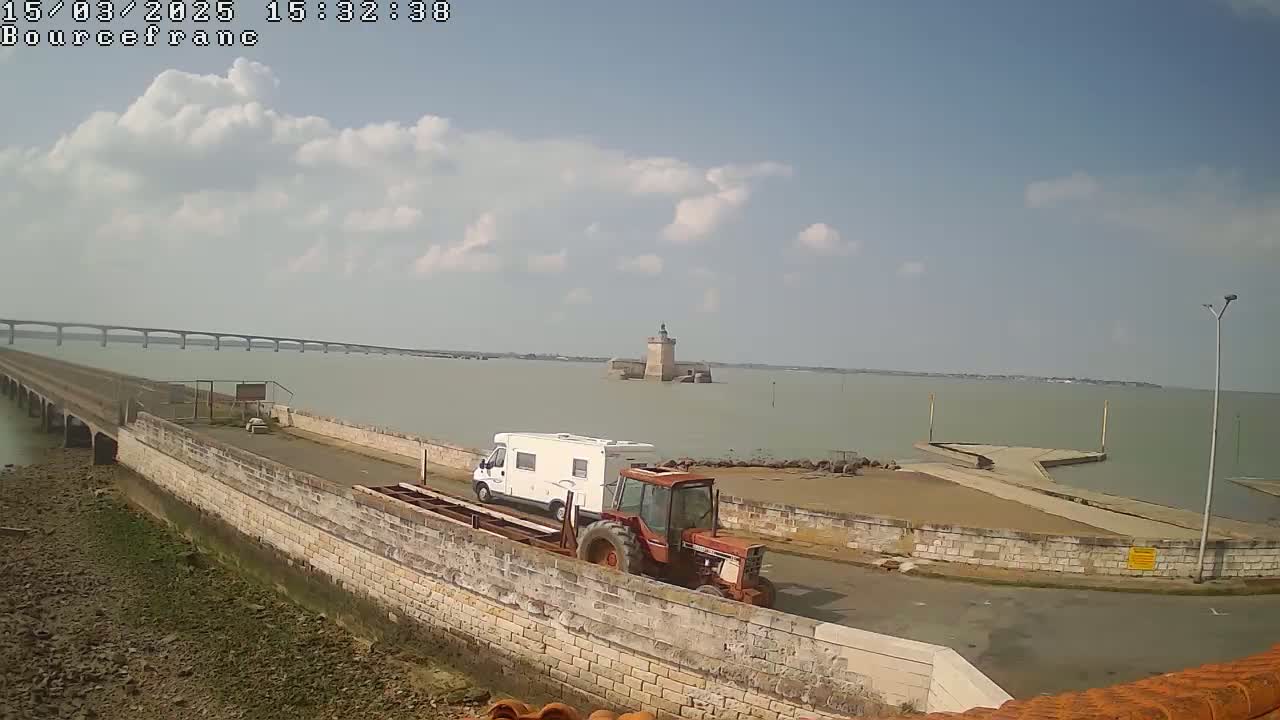 A white campervan and a red tractor are parked on a stone causeway leading to a small island with a tower, under a partly cloudy sky.