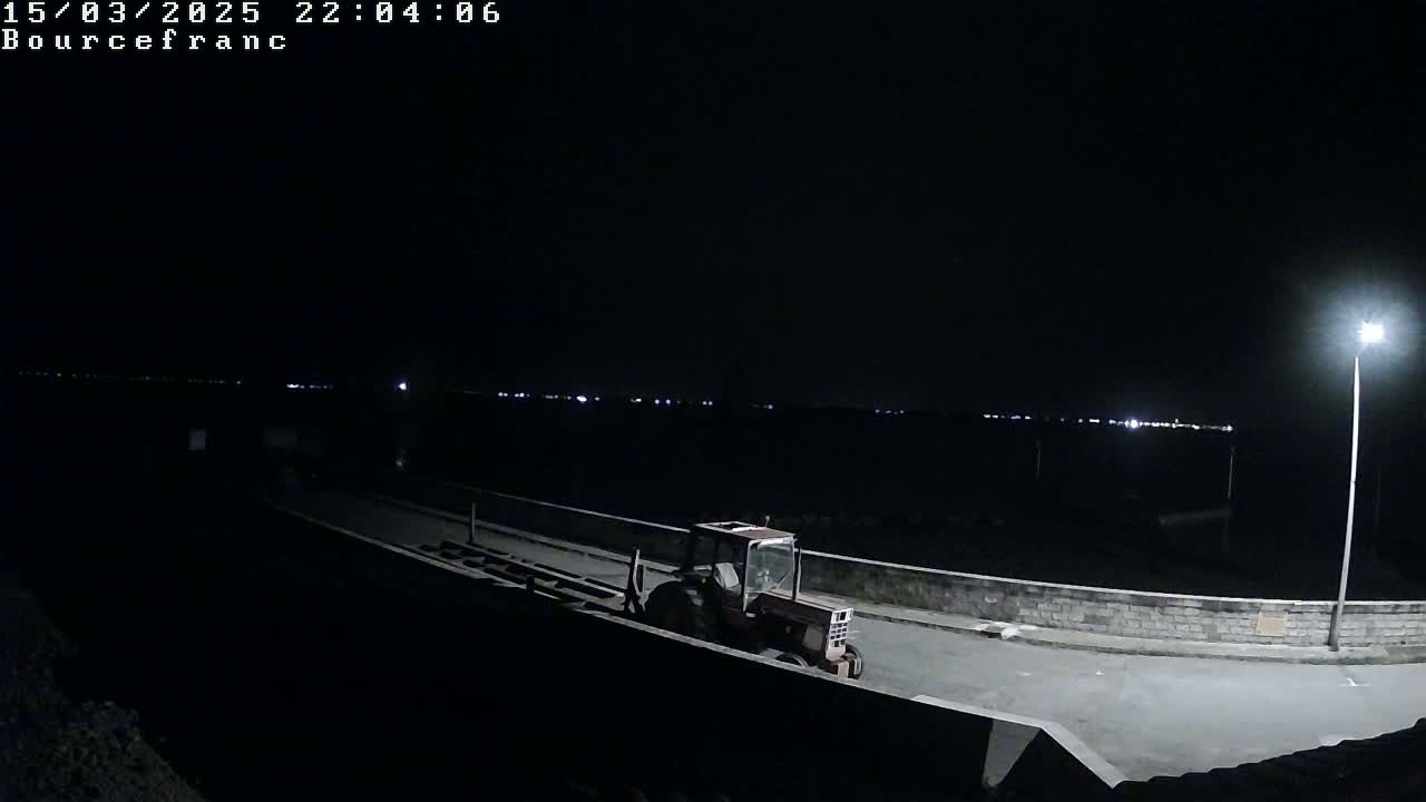 A tractor sits on a dark, paved area next to a low stone wall at night, with distant lights visible in the background.