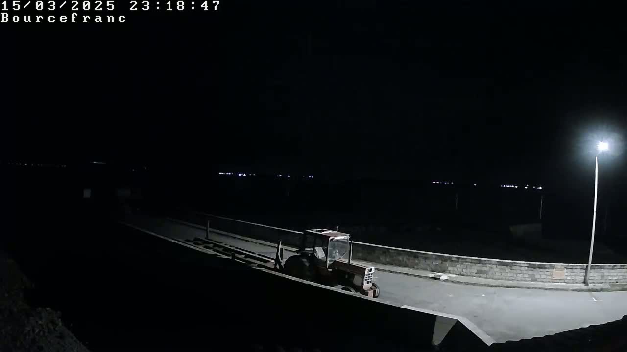 A red tractor is parked on a dark road at night, illuminated by a single streetlight, with a distant horizon of faint lights.